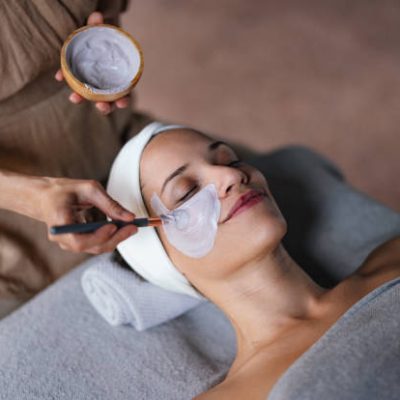 Close up shot of a beautiful smiling woman lying with her eyes closed while another woman is applying a face mask on her in the spa center. She is relaxed and enjoys the treatment.
